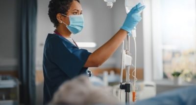 Hospital Ward: Professional Black Head Nurse Wearing Face Mask Does Checkup of Patient's Vitals, Checking Heart Rate Computer, Intravenous or Iv Fluids Drip Bag. Caring Nurse Monitors Person Recovery
